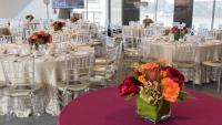 A large event space with floor to ceiling windows, tables with ivory tablecloths, and flower arrangements